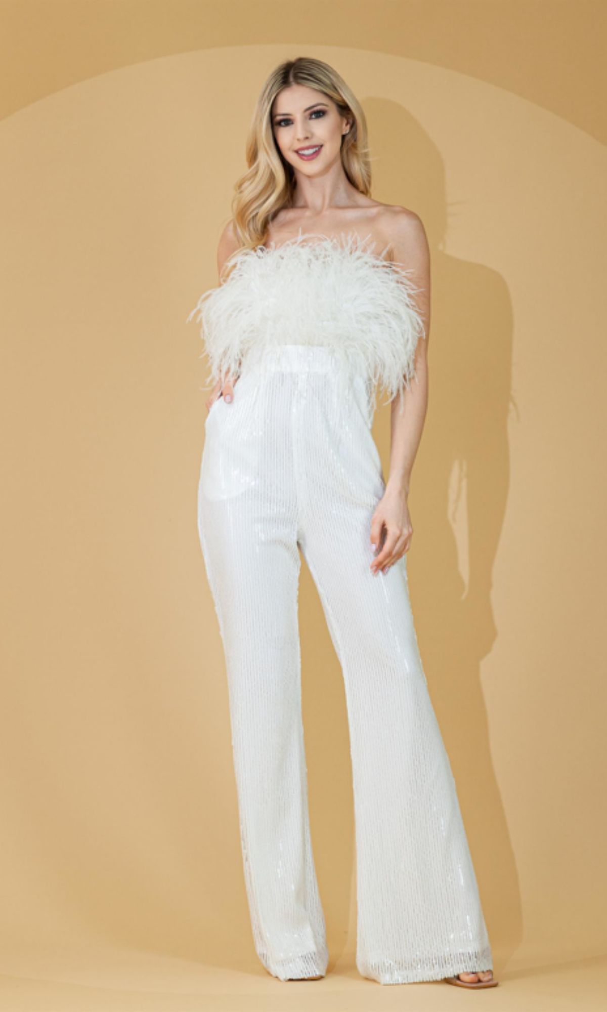 Woman poses against a beige background, smiling with hands in pockets, wearing the White Feather-Trimmed Long Sequin Prom Jumpsuit 3019 featuring a strapless design and feathered bodice trim.